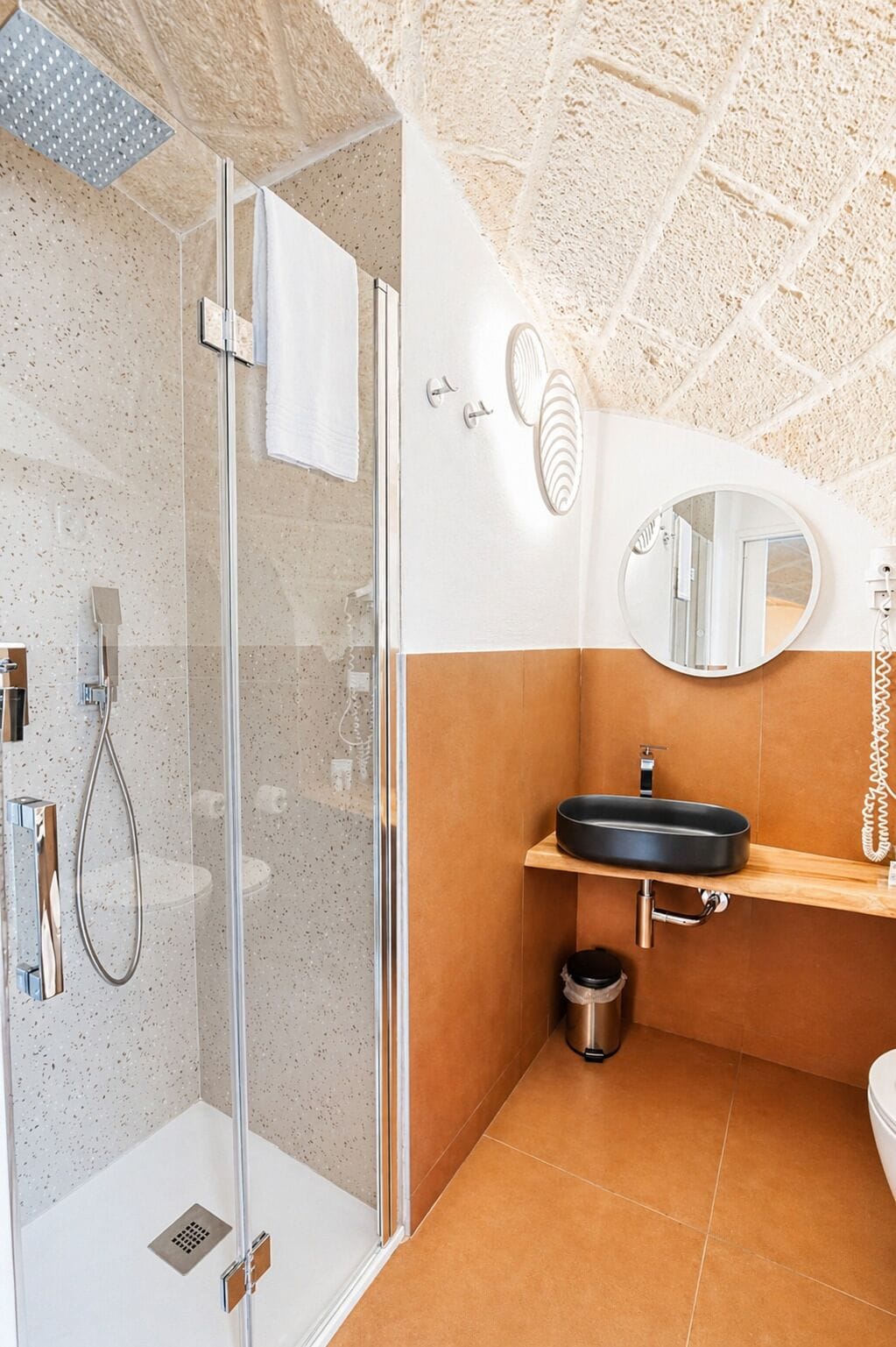 A modern bathroom with a glass-enclosed shower, a round mirror, and a black sink on a wooden shelf.