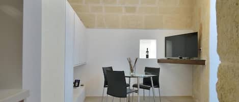 A compact dining area with a glass table and four chairs, a flat-screen TV mounted on a wall, and a stone wall above.