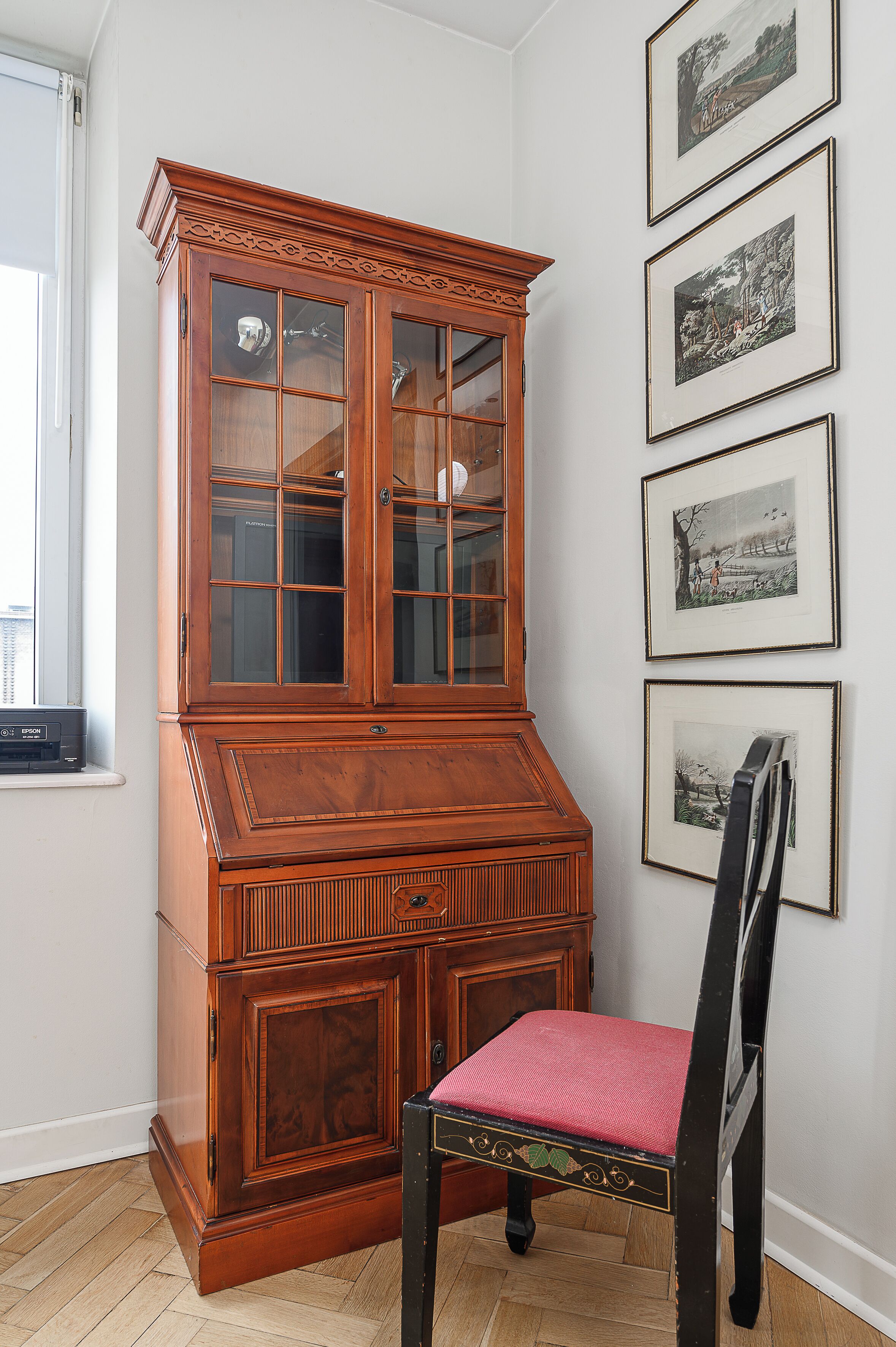 A wooden cabinet with glass doors, a chair with a red cushion, and framed pictures on the wall.