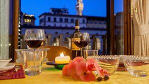 Uma mesa posta com copos de vinho, uma vela e objetos decorativos, com vista para um edifício e uma torre ao fundo.