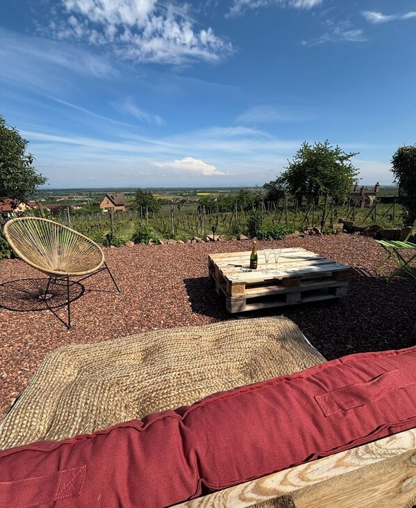 A patio with a wicker chair, a wooden table with a bottle and glass, and a red cushion.