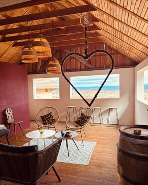 A cozy room with wicker furniture, a heart-shaped decoration, and large windows offering a view of the beach.