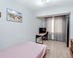 Um quarto com cama, escrivaninha com cadeira, televisão e janela com cortinas.
