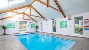 Una piscina interior con agua cristalina azul, vigas de madera en el techo y ventanas amplias.