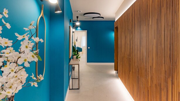 Un pasillo moderno con paredes azules, paneles de madera en un lateral y un espejo que refleja una habitación con una planta y una silla.