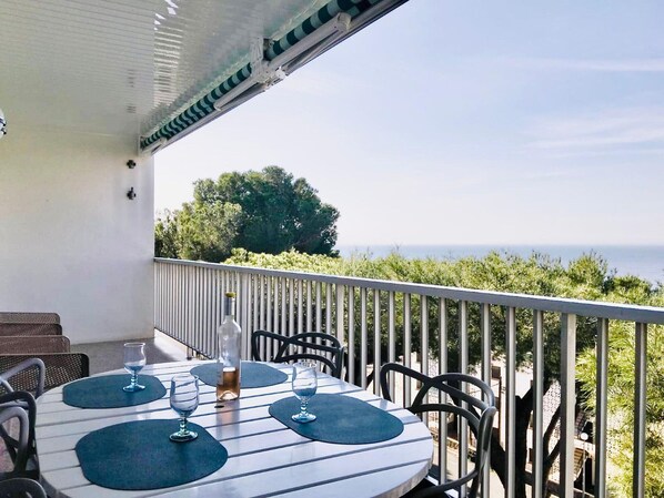 A round table with four chairs and a bottle of wine, set on a balcony with a railing, overlooking a coastal landscape.