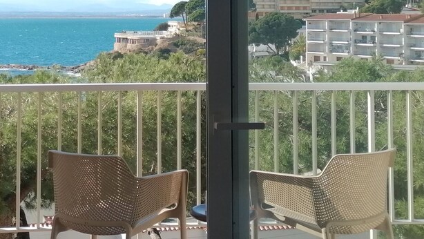 Un balcon agrémenté de deux fauteuils en osier, offrant une vue sur le paysage urbain de la côte, avec ses bâtiments, et une vue dégagée sur la mer.