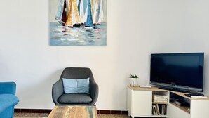 Un salon avec un canapé moderne, une table basse, un téléviseur à écran plat, un meuble de rangement blanc et un tableau représentant des voiliers.