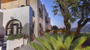 Un edificio moderno con ventanales arqueados y un balcón, rodeado de abundante vegetación y vistas a montañas lejanas.