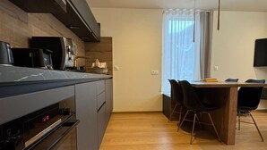 Moderni keittiö, jossa on sisäänrakennettu uuni ja mikroaaltouuni, ruokailutila puupöydällä ja mustilla tuoleilla sekä suuri ikkuna, jossa on läpikuultavat verhot.