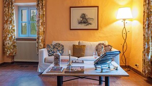 A cozy living room with a sofa, a coffee table with books and a candle, a framed picture on the wall, and a lamp. - La Foresteria Castell'In Villa (Castelnuovo Berardenga)