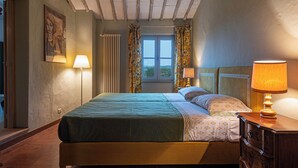 A bed with a green blanket, two bedside lamps, a window with curtains, and a radiator. - La Foresteria Castell'In Villa (Castelnuovo Berardenga)
