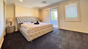 A bedroom with a tufted headboard, a bed with a floral bedspread, a nightstand with a lamp, and a sliding glass door leading to a balcony. - Family and Group Holiday Home Sleeps 13 Near Western Sydney Airport, Penrith (Jordan Springs)