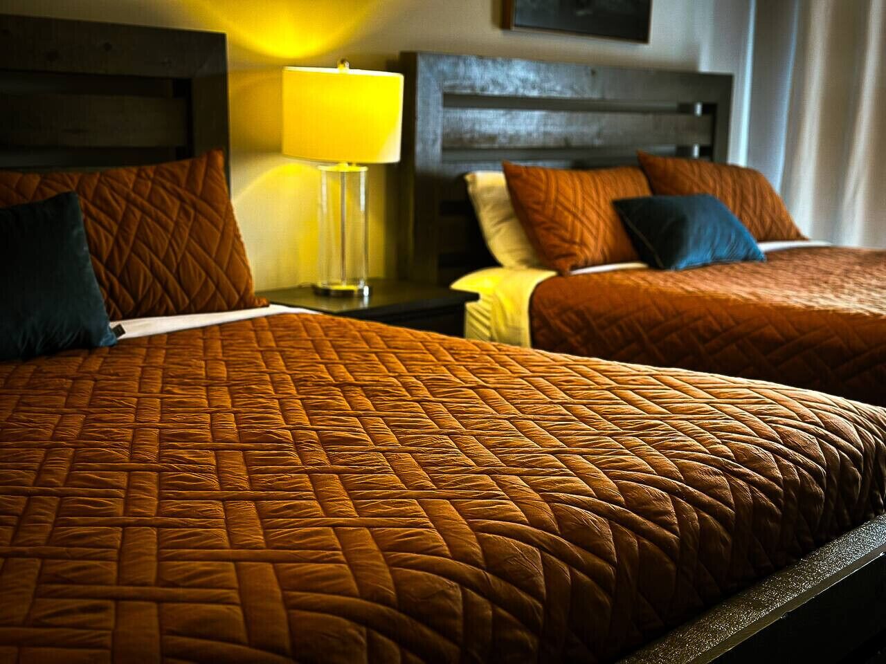 A hotel room with two beds, each with a quilted bedspread and pillows. There is a nightstand with a lamp between the beds.