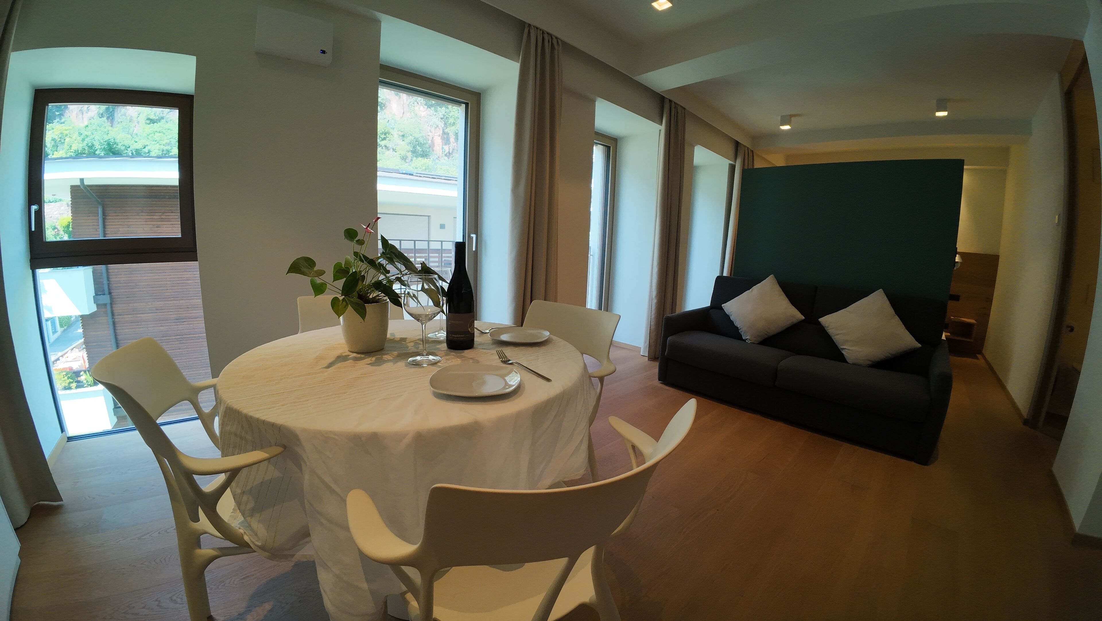 A dining area with a round table, chairs, a potted plant, and a bottle of wine. There is a dark-colored sofa and a green accent wall in the living area.