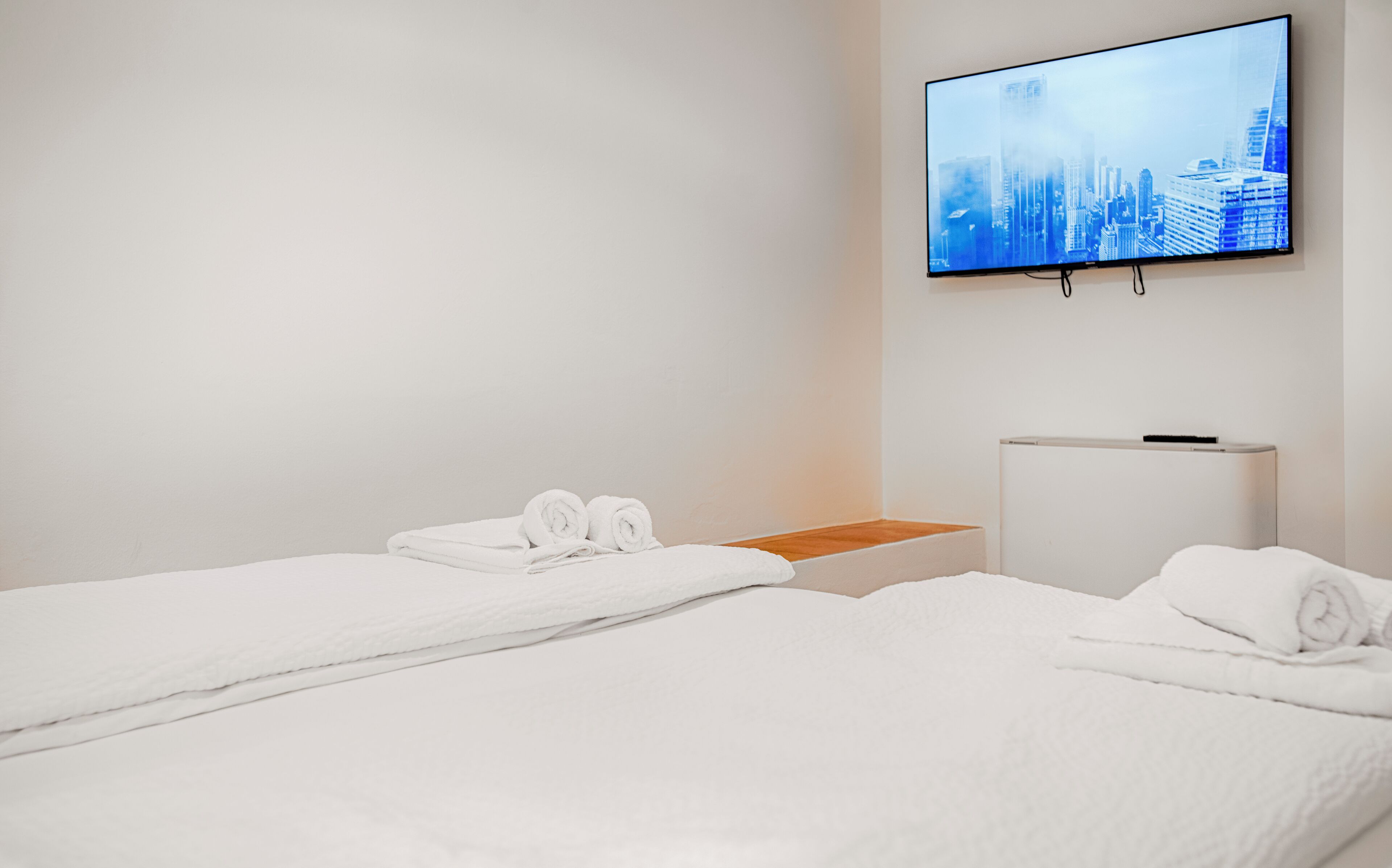 Quarto de hotel com duas camas, uma TV fixada na parede e um pequeno armário branco.