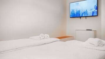 Quarto de hotel com duas camas, uma TV fixada na parede e um pequeno armário branco.