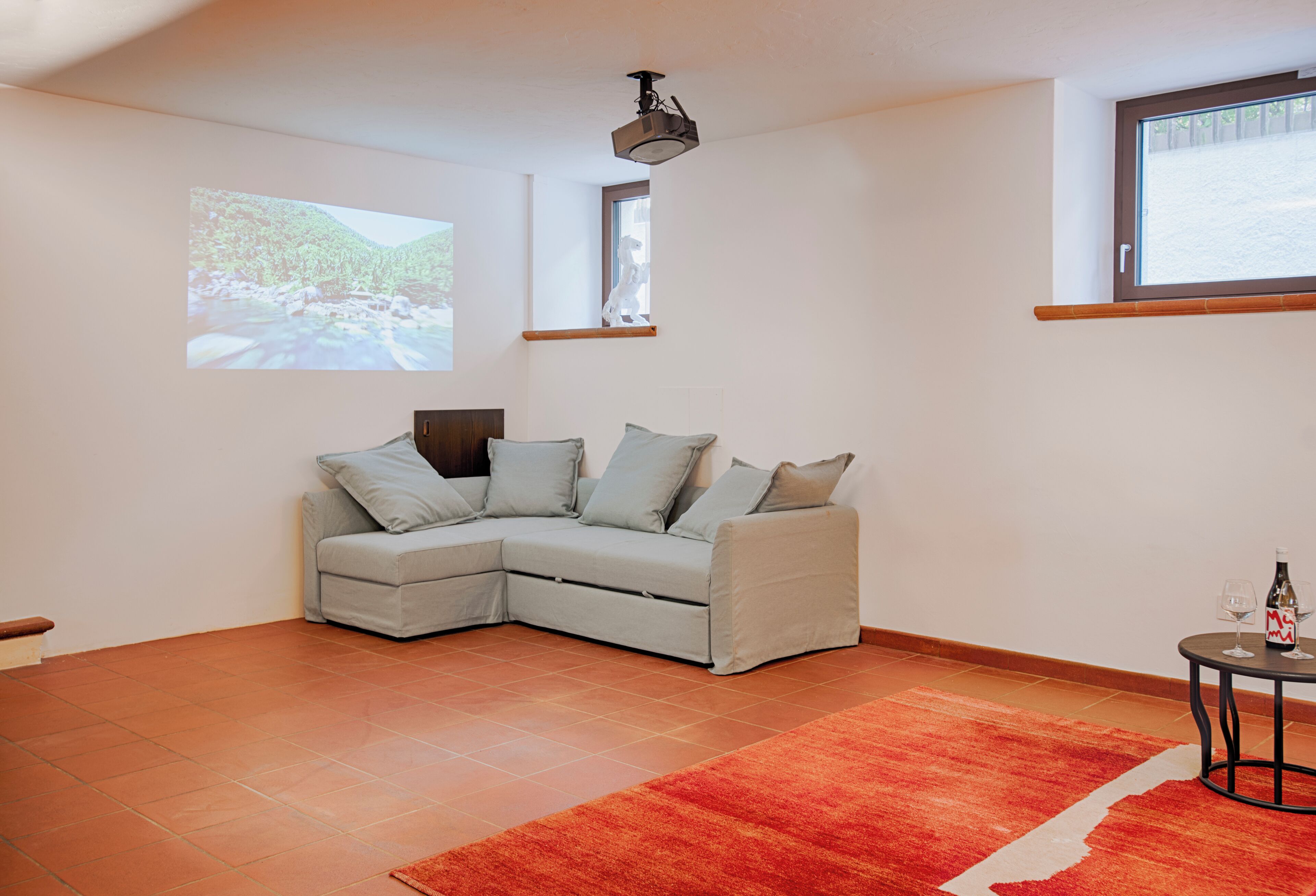 Un salon moderne avec un canapé d’angle, un tapis rouge, un projecteur au plafond et une fenêtre donnant sur des arbres.