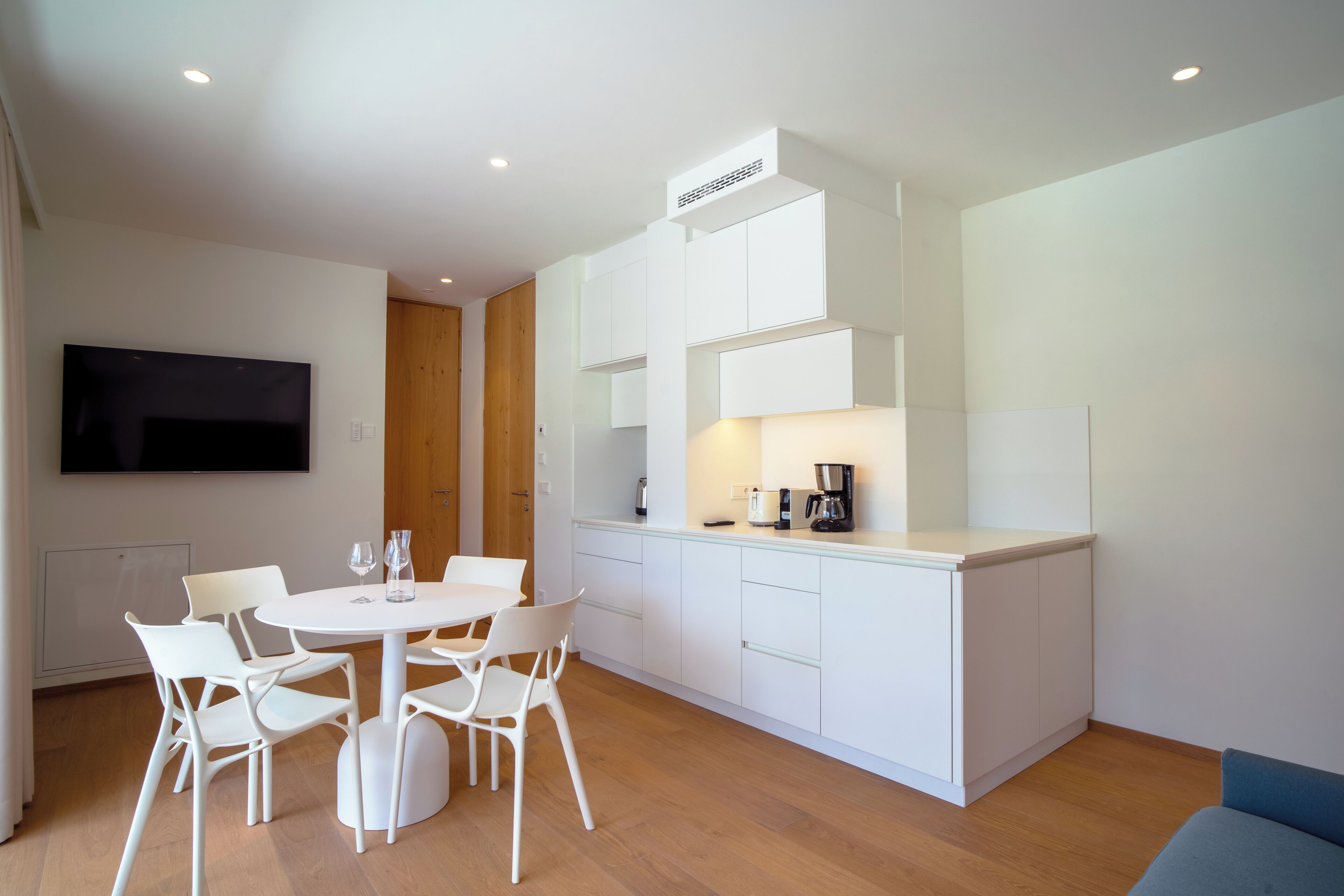 A modern, minimalist kitchen with white cabinetry, a built-in stove, and a dining area with white chairs and a round table.