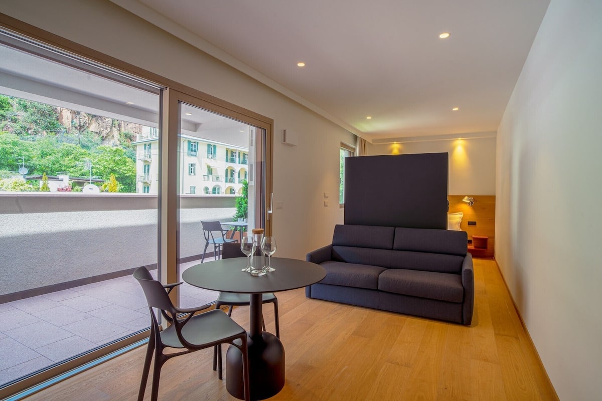 A modern hotel room with a balcony, a round table with chairs, a sofa, and a view of buildings and greenery outside.