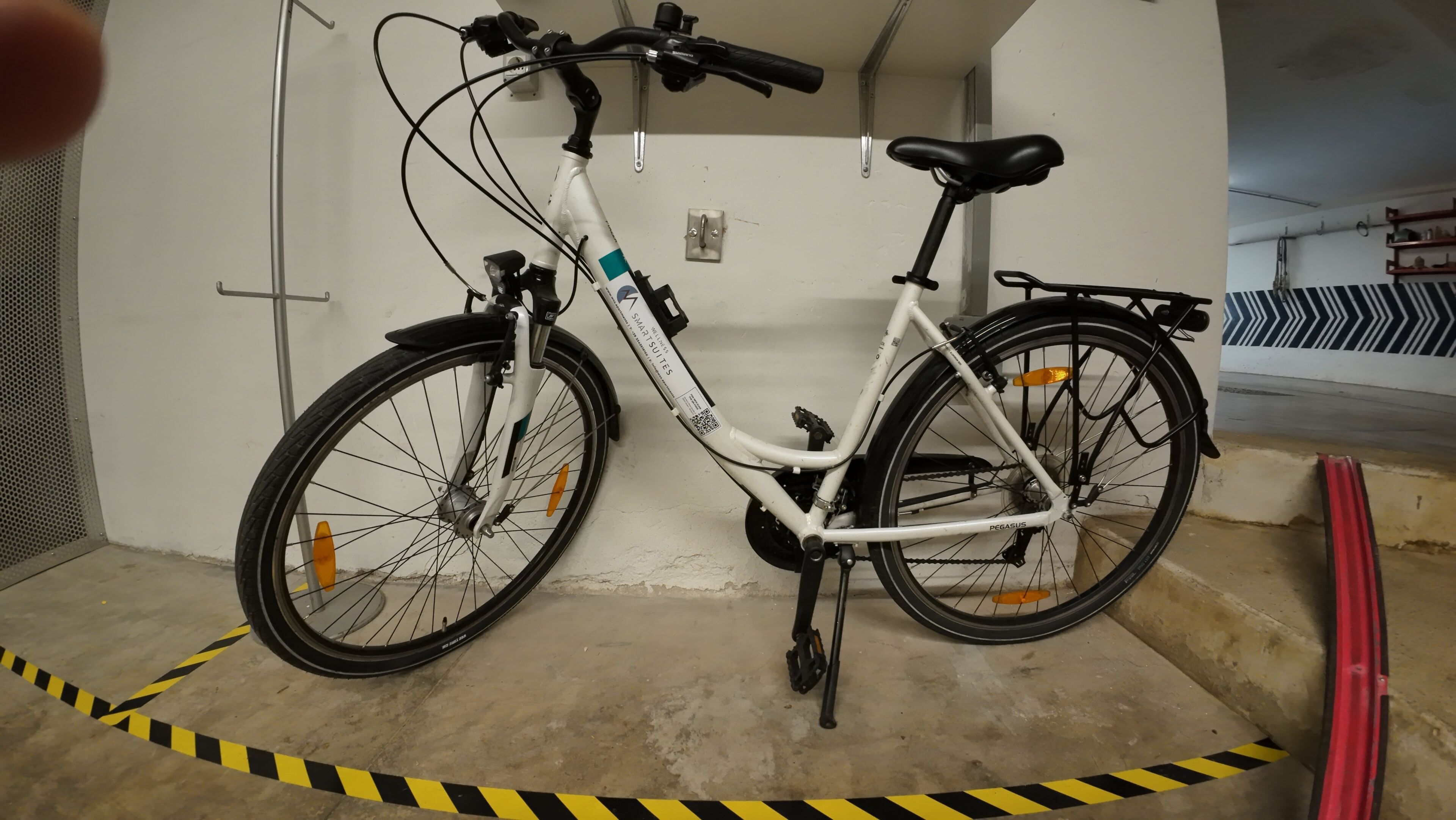 Un vélo blanc avec une selle et un guidon noirs, garé dans un garage où du ruban de signalisation jaune et noir est disposé sur le sol.