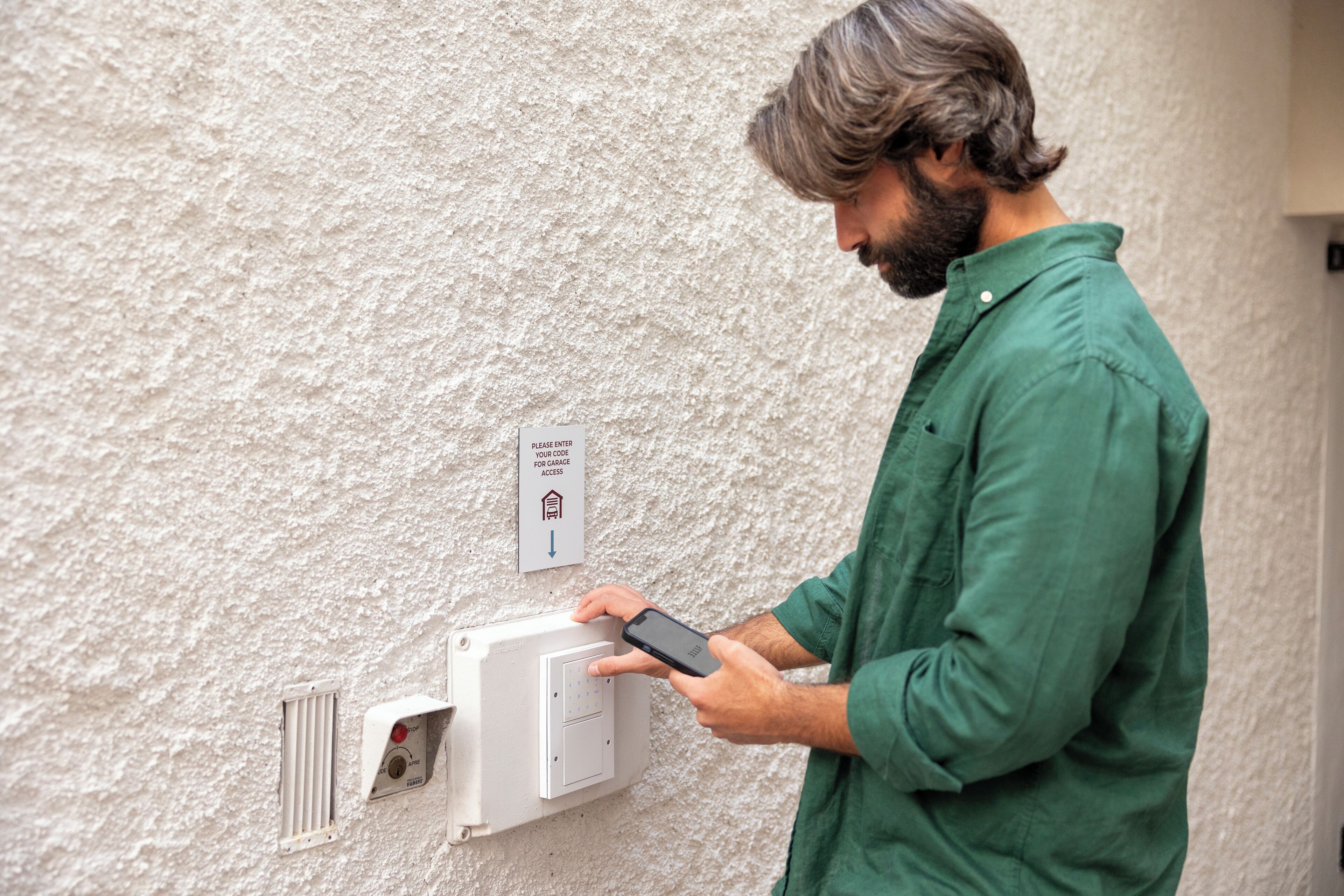 A person using a smartphone to interact with a wall-mounted device, possibly for access control, with a sign indicating a code entry requirement.