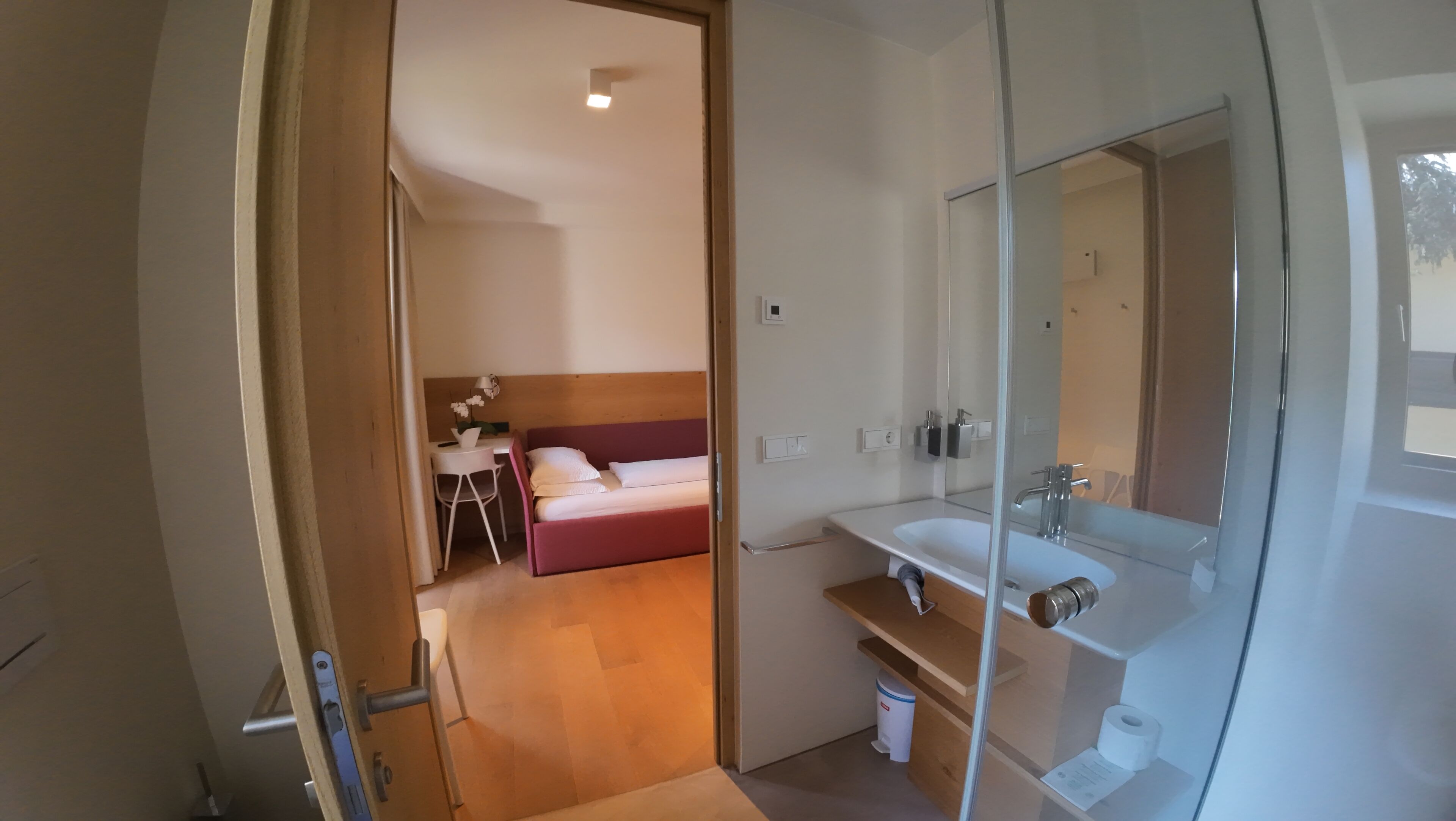 A hotel room with a bed, a desk with a chair, a bathroom with a sink and mirror, and a small trash bin.