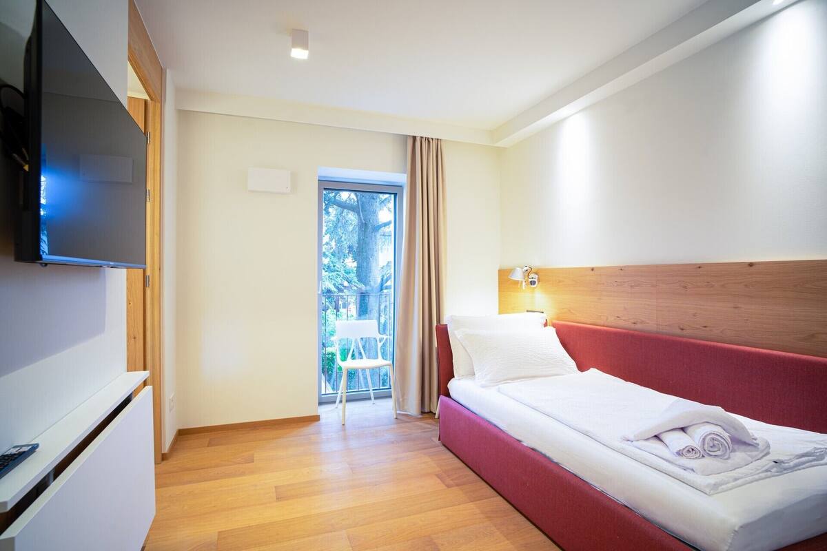 A hotel room with a bed, a TV, a wooden floor, and a balcony with a chair.