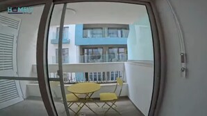 A balcony with a yellow table and chairs, a glass railing, and a view of a building with blue and white exterior. - Centrally located in Qawra, this modern 2-bed apartment offers comfort, style & easy access to the promenade — perfect for a carefree holiday escape! (San Pawl il-Baħar)