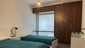 A bedroom with two beds, a desk, a chair, a wardrobe, and a window with blinds. - Centrally located in Qawra, this modern 2-bed apartment offers comfort, style & easy access to the promenade — perfect for a carefree holiday escape! (San Pawl il-Baħar)