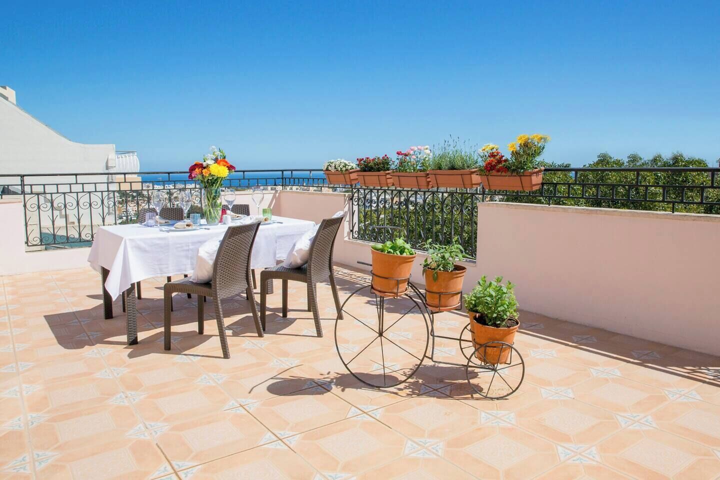 A terrace with a table set for two, surrounded by potted plants and a clear view of the sea.