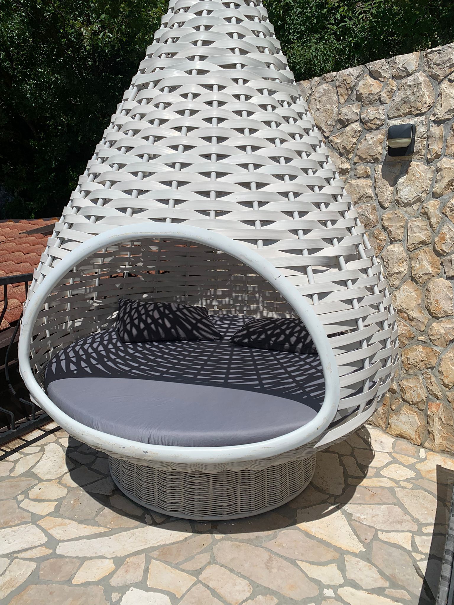 A round, white, woven outdoor seating area with a cushioned seat and backrest, set against a stone wall.