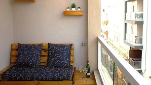 A balcony with a wooden bench, two cushions, a small table with a bottle and glass, and potted plants on the wall.