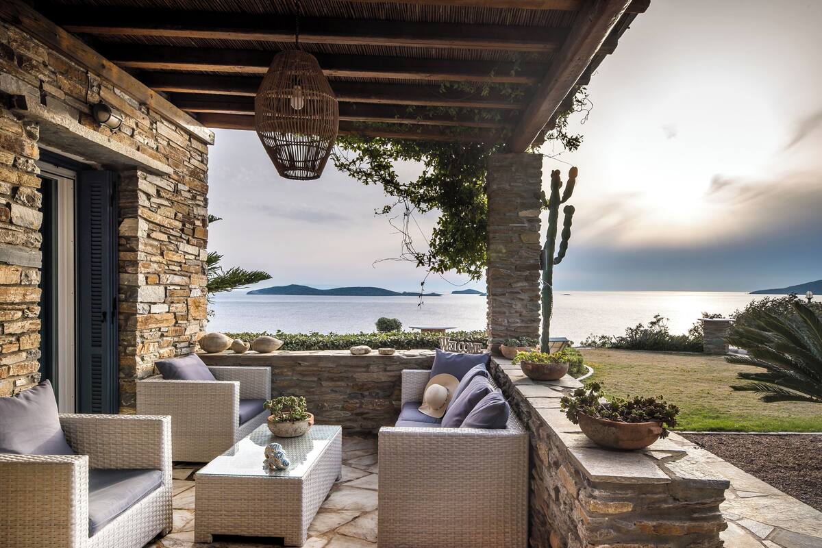A stone-walled outdoor seating area with wicker furniture, a welcome sign, and a view of the sea.