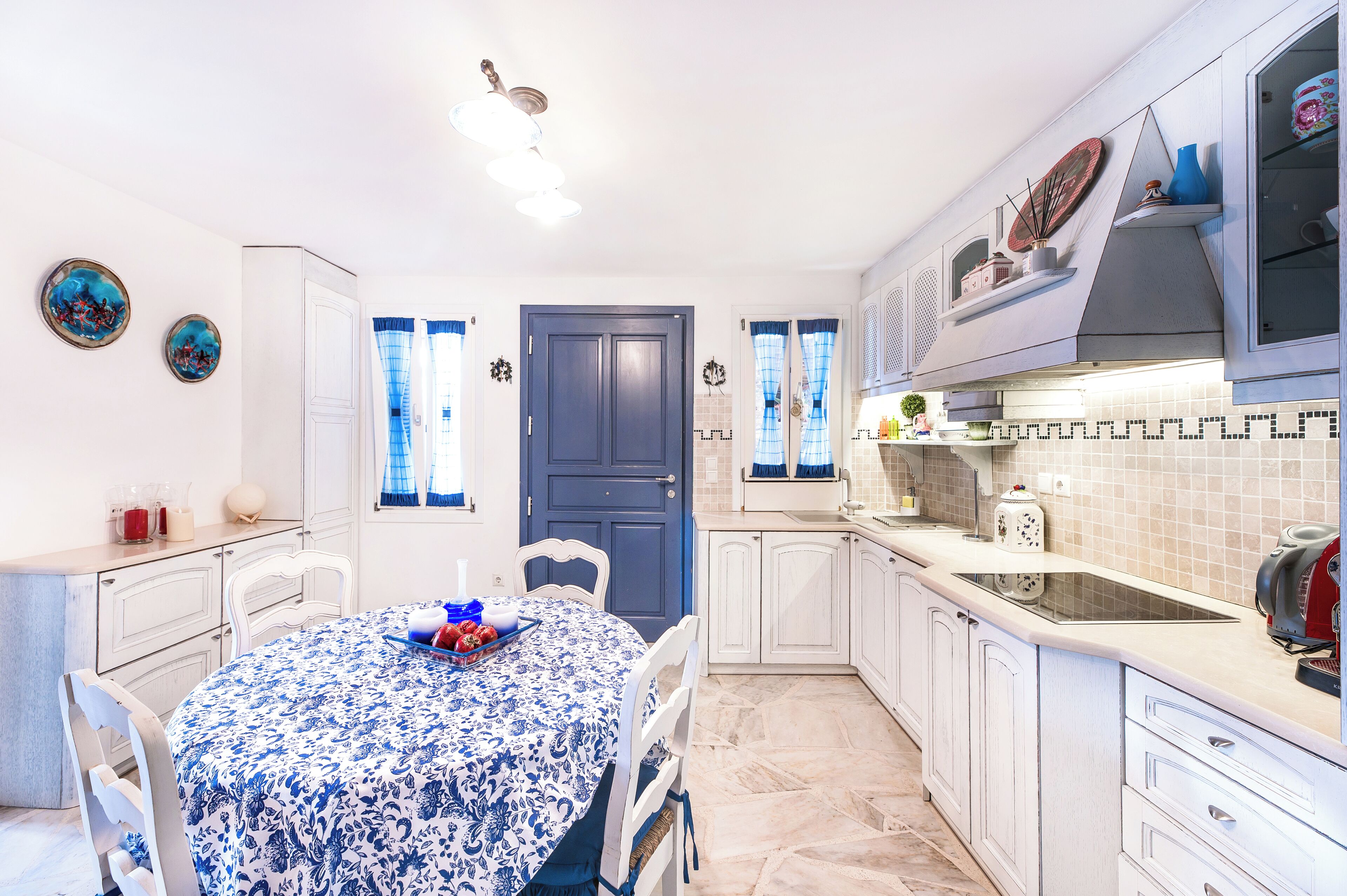 A modern kitchen with white cabinetry, a blue door, and a dining area with a blue and white tablecloth.
