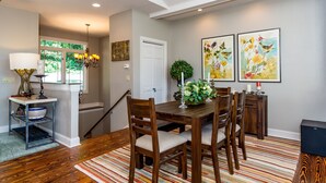 A dining area with a wooden table, chairs, and a striped rug. There is a staircase leading to another level, a window with a view of greenery, and a door.