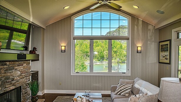 A living room with a stone fireplace, a large window with a view of a green landscape, and a ceiling fan.