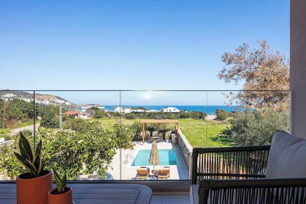 Un balcón con vistas a una piscina y al mar, un sofá de mimbre y plantas en macetas.