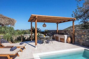 Un área junto a la piscina con sillones reclinables, una pérgola, una mesa con sillas y un muro de piedra.