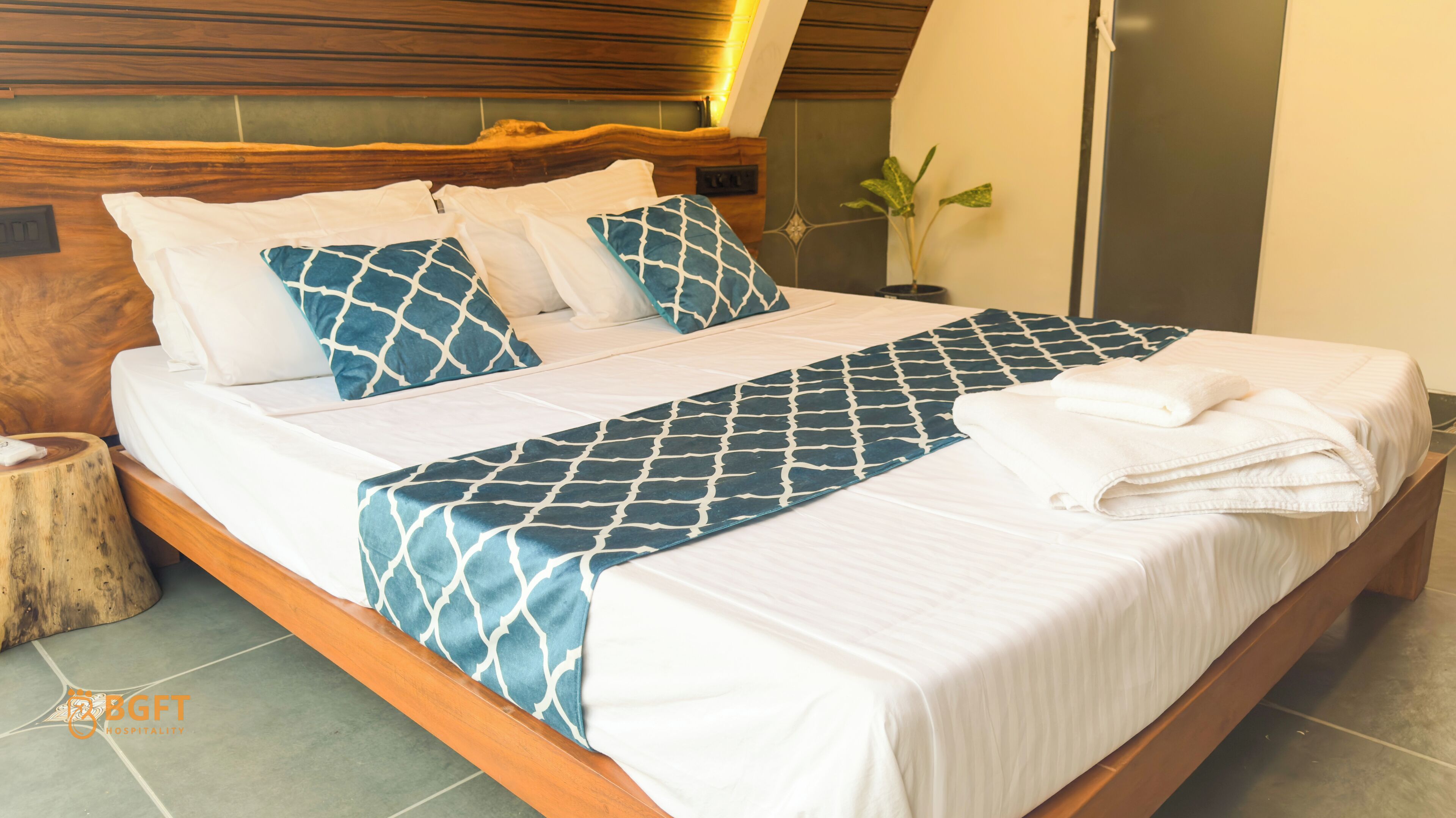 A neatly made bed with a patterned bedspread and pillows, a wooden headboard, and a bedside plant.