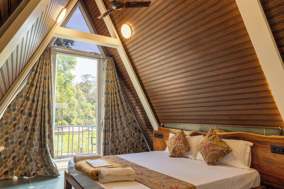 Ein Schlafzimmer mit schrägen Wänden, einem Bett mit Kissen, einem Nachttisch und einem Fenster mit Vorhängen.