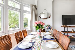 A dining area with a table set for four, a sofa, a TV, and a window with curtains. - Mill Hill Duplex | 3BR · Balcony · London in 28 Mins (London)