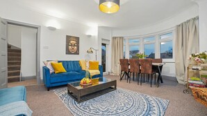 Ein Wohnzimmer mit blauem Sofa, einem Couchtisch mit Früchten, einem Essbereich mit Stühlen und einem Fenster mit Vorhängen.