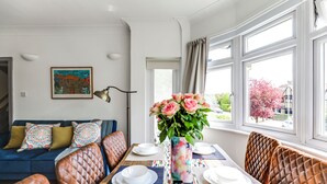 Ein Essbereich mit einem Tisch, der für vier Personen gedeckt ist, einer Couch mit gemusterten Kissen, einer Vase mit Blumen und einem Bild an der Wand.