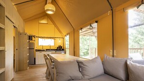 A cozy, open-plan living area with a wooden deck, a sofa with cushions, a kitchen area with a refrigerator and a window overlooking greenery.
