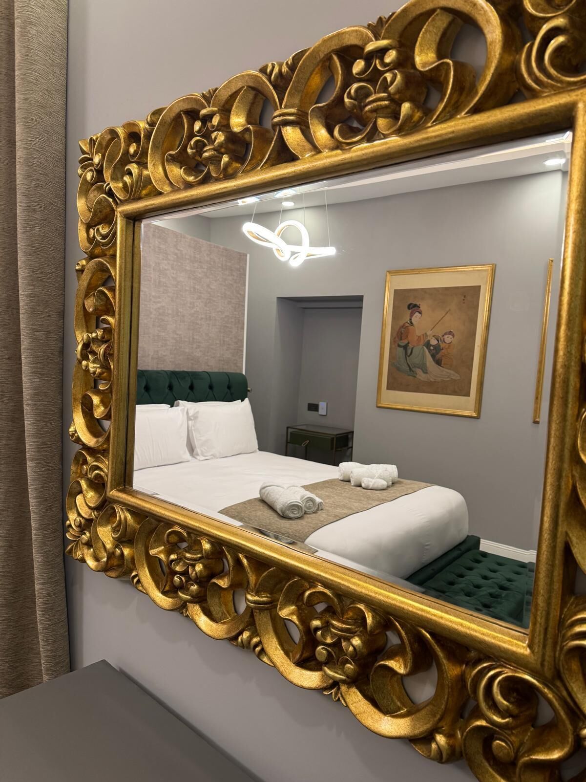 A hotel room with a large ornate gold-framed mirror, a neatly made bed with white linens, and a green upholstered headboard.