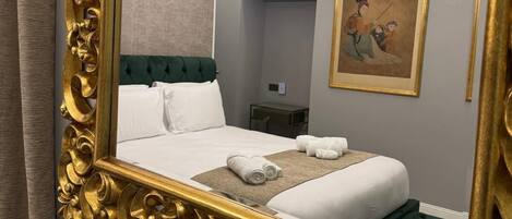 A hotel room with a large ornate gold-framed mirror, a neatly made bed with white linens, and a green upholstered headboard.