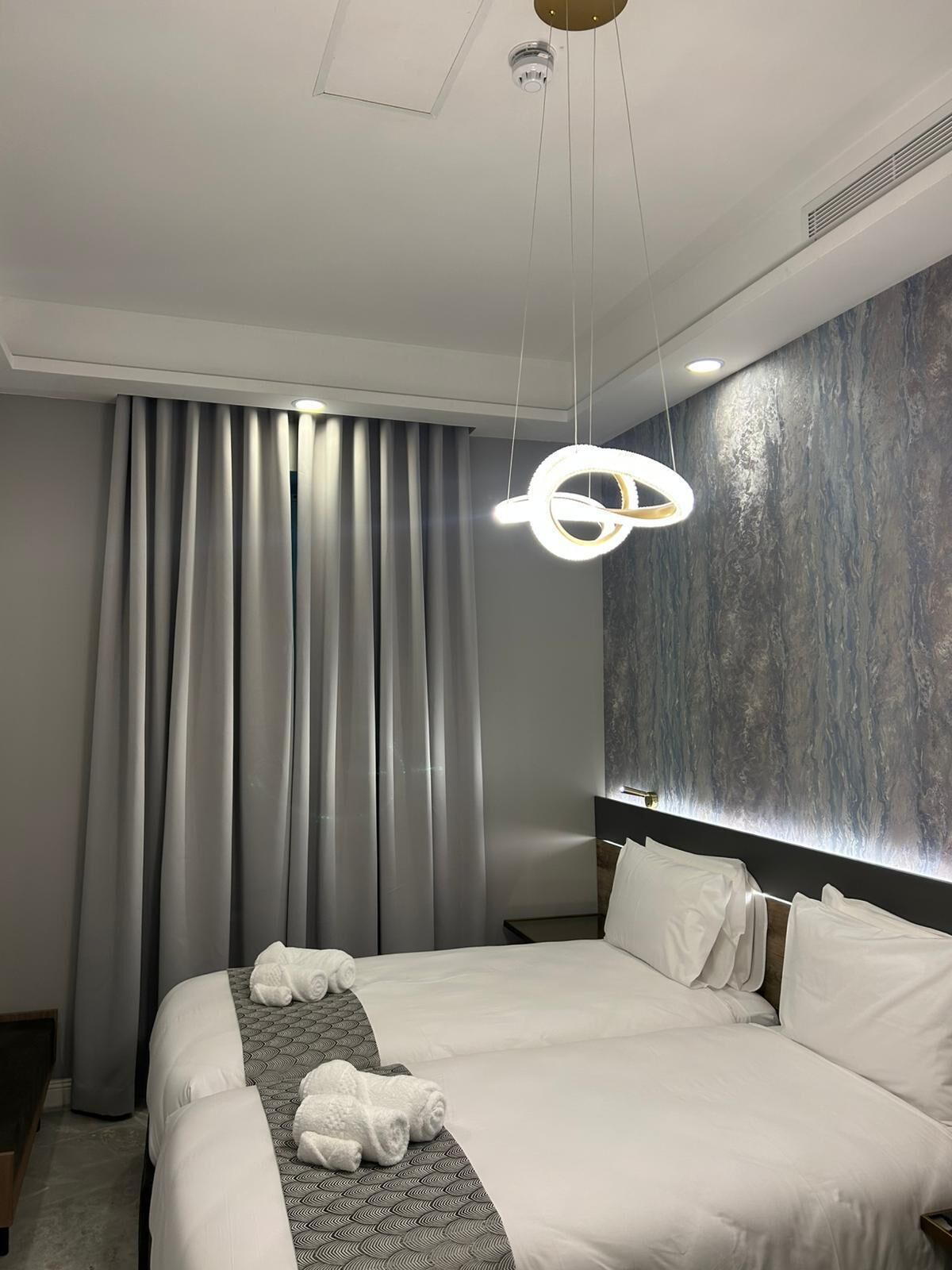 A modern hotel room with a large bed, grey curtains, a unique circular light fixture, and a textured wall.