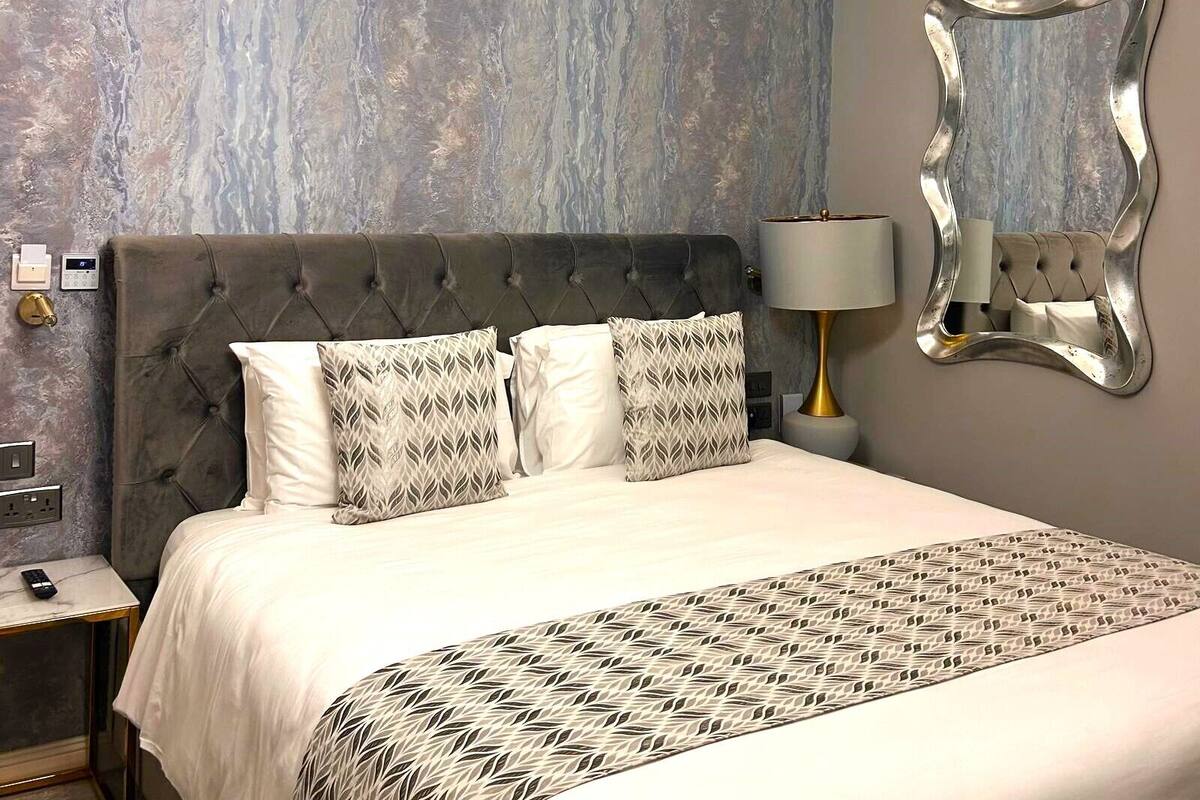 A bedroom with a tufted headboard, a patterned bedspread, a modern light fixture, a wall-mounted mirror, and a bedside lamp.