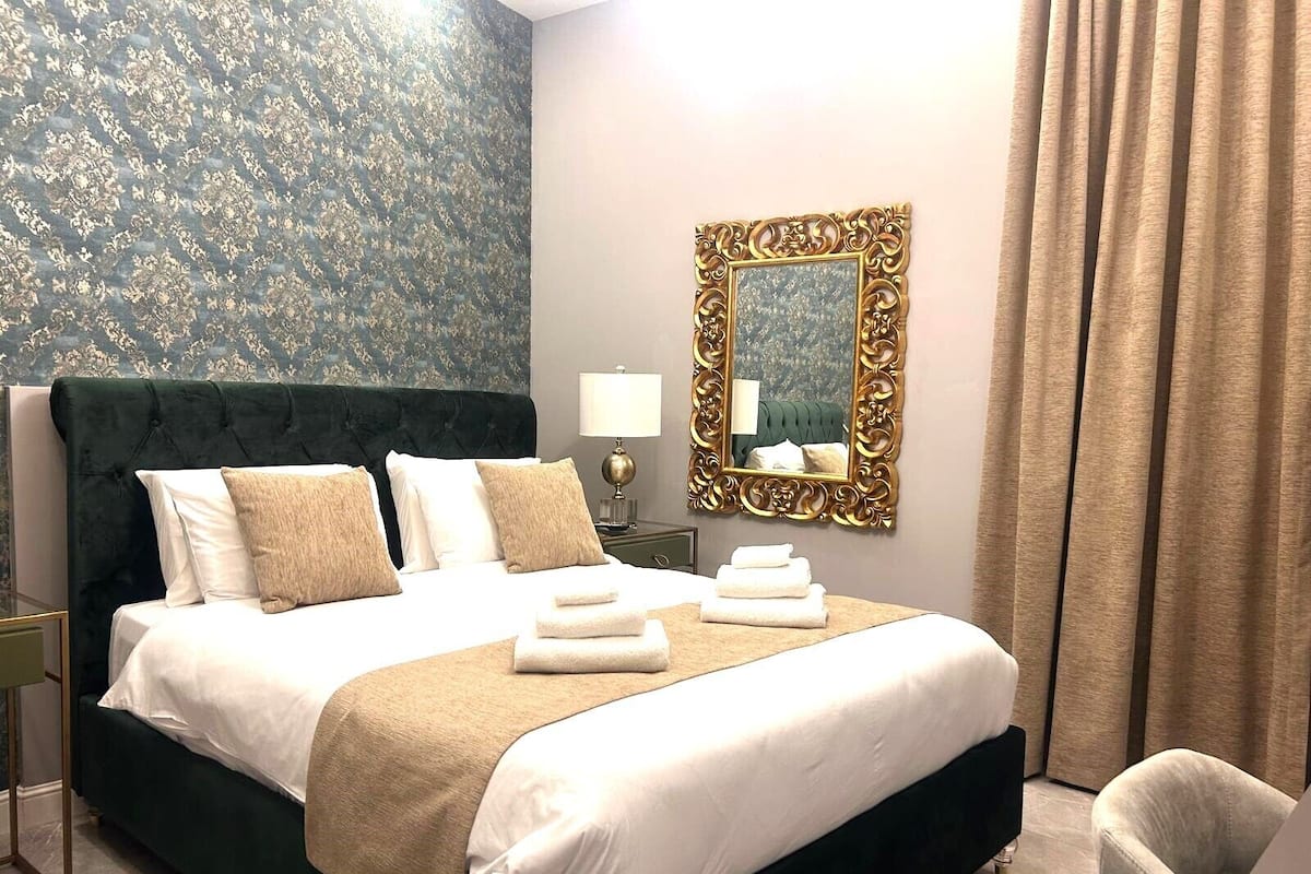 A neatly made bed with white and beige linens, a dark green upholstered headboard, and a gold-framed mirror on the wall.